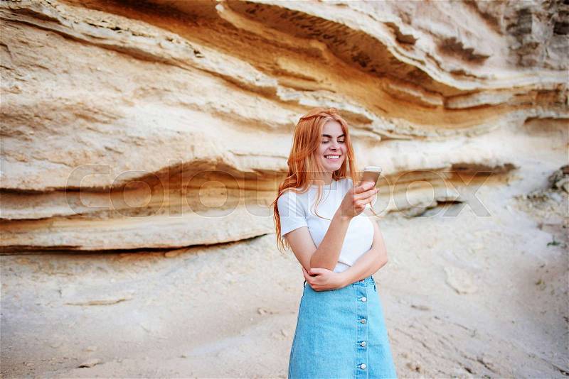 Beautiful woman smiling and looking at the screen of a smartphone on the background of a sand quarry, stock photo