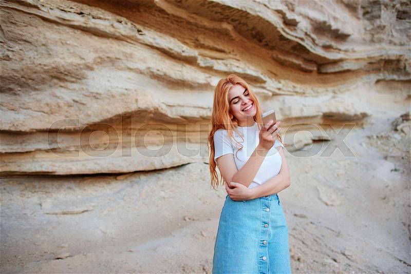 Beautiful woman smiling and looking at the screen of a smartphone on the background of a sand quarry, stock photo