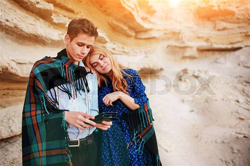 Beautiful woman and handsome man wrapped in a blanket. They are smiling and looking at the screen of a tablet on the background of a sand quarry, stock photo