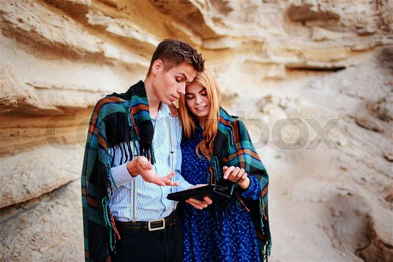 Beautiful woman and handsome man wrapped in a blanket. They are smiling and looking at the screen of a tablet on the background of a sand quarry, stock photo