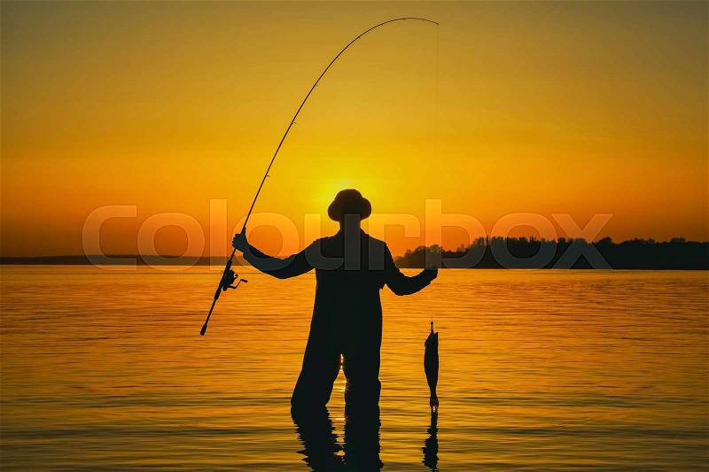 A fisherman with a fishing rod in his hand and a fish caught stands in the water against a beautiful sunset, stock photo