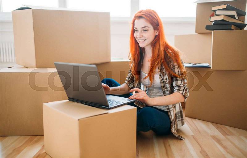 Smiling girl using laptop among cardboard boxes, moving to new house, housewarming, stock photo