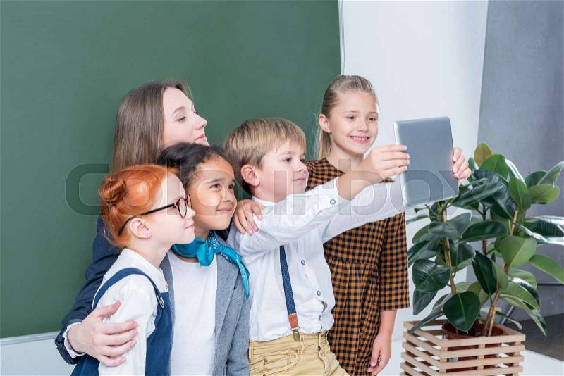 Young female teacher with cute little students taking selfie with digital tablet in class, stock photo