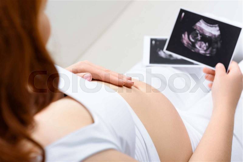 Cropped view of pregnant woman touching her belly and holding ultrasound scans at home, stock photo