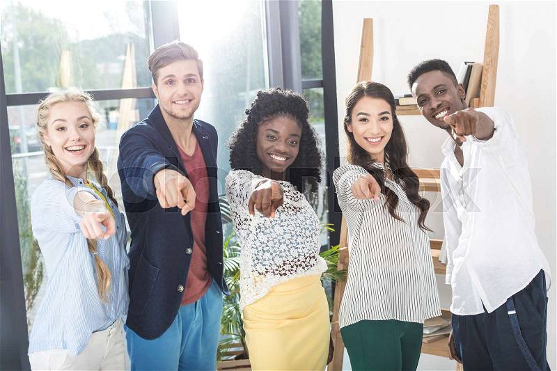 Multiethnic smiling business people pointing at camera while standing together in office, stock photo