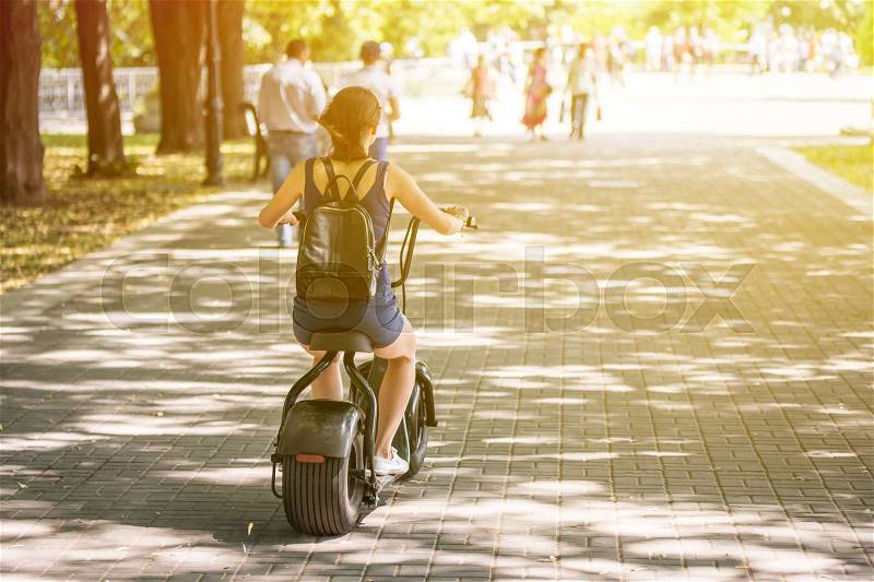 A young woman riding zero-emission eco electric scooter bike in a city park. Back view. Day, sunlight. , stock photo