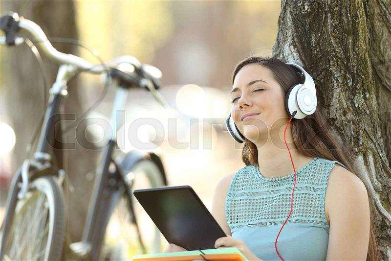 Single student wearing headphones listening to music on line with a tablet outdoors in a park, stock photo