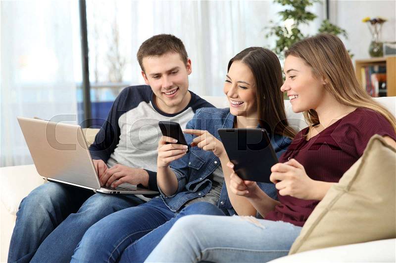Three happy friends sharing on line content with multiple devices sitting on a sofa at home, stock photo