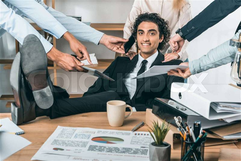 Portrait of smiling busy businessman sitting at workplace while colleagues helping with work in office, stock photo