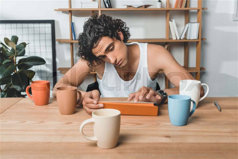 Portrait of tired businessman in underwear using tablet while sitting at workplace with cups of coffee in office, stock photo