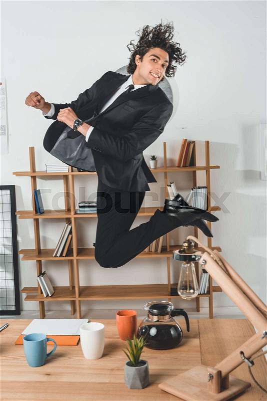 Smiling businessman jumping near workplace with coffee pot and tablet in office, stock photo