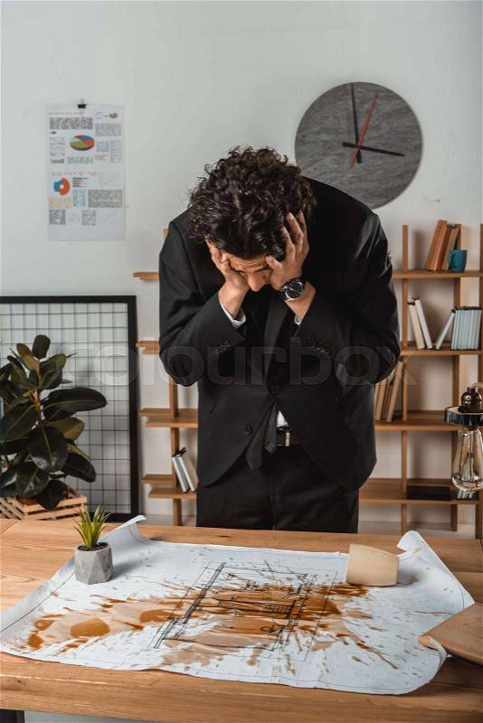 Sad businessman looking at spoiled blueprints with coffee on it at workplace, stock photo