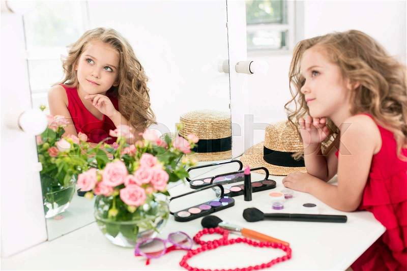 A little girl with red dress and cosmetics. She is in mother\'s bedroom, sitting and looking at mirror, stock photo