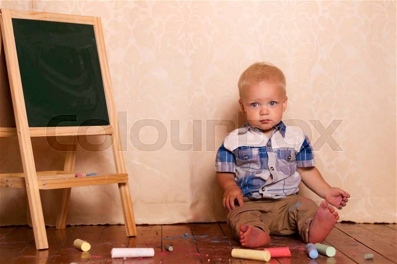 Adorable little boy sitting near drawing board. Cute toddler among chalk for drawing and looking at camera, stock photo