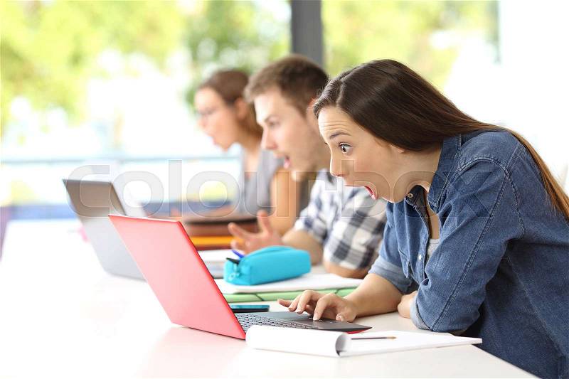 Side view of three amazed students receiving good news on line in a classroom, stock photo