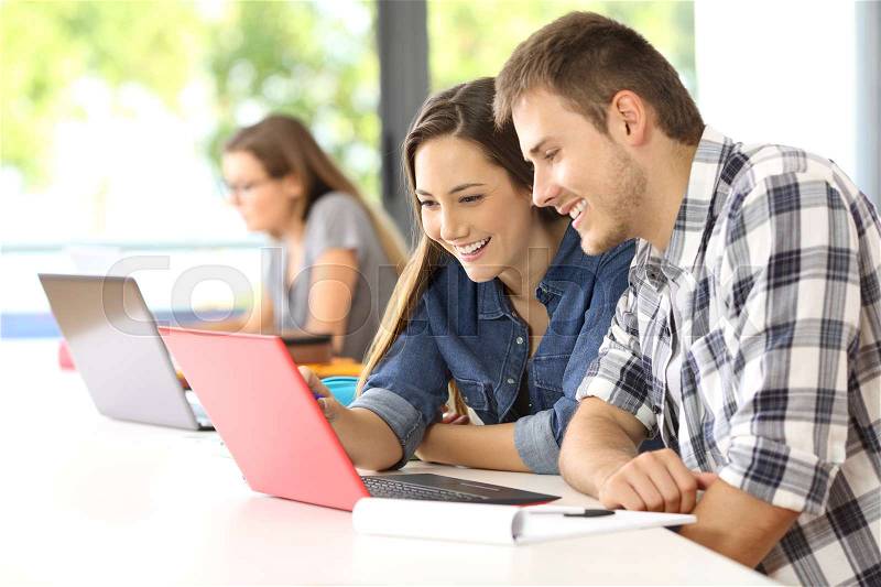Two happy students learning on line together with a computer in a classroom with other classmate in the background, stock photo