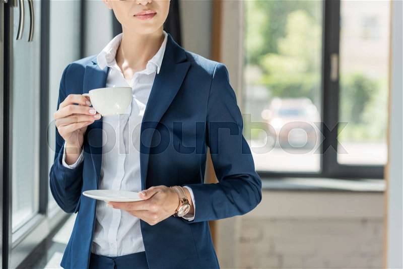 Cropped view of businesswoman in suit drinking coffee in office, stock photo