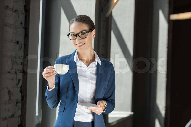 Beautiful smiling businesswoman in suit drinking coffee in office, stock photo