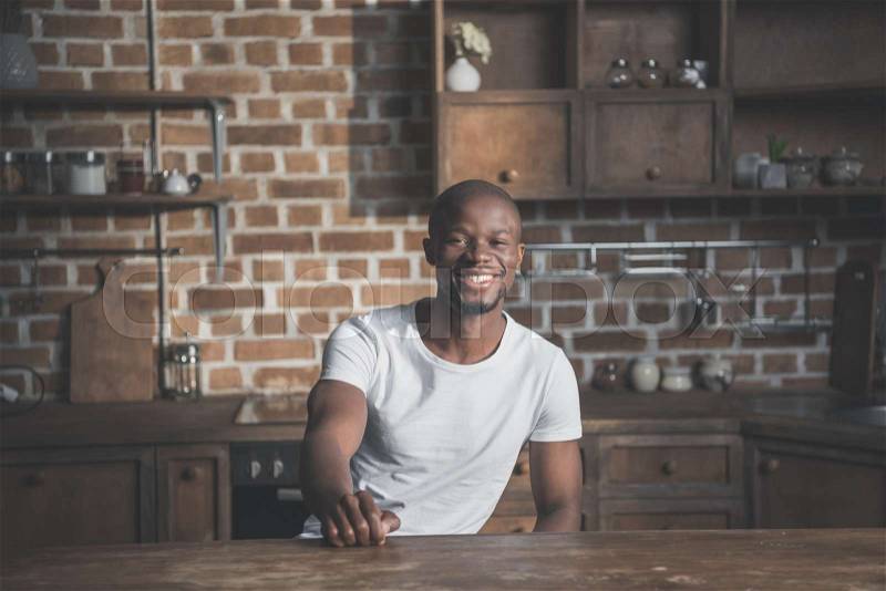 Portrait of young happy handsome african american man sitting at table in kitchen, stock photo