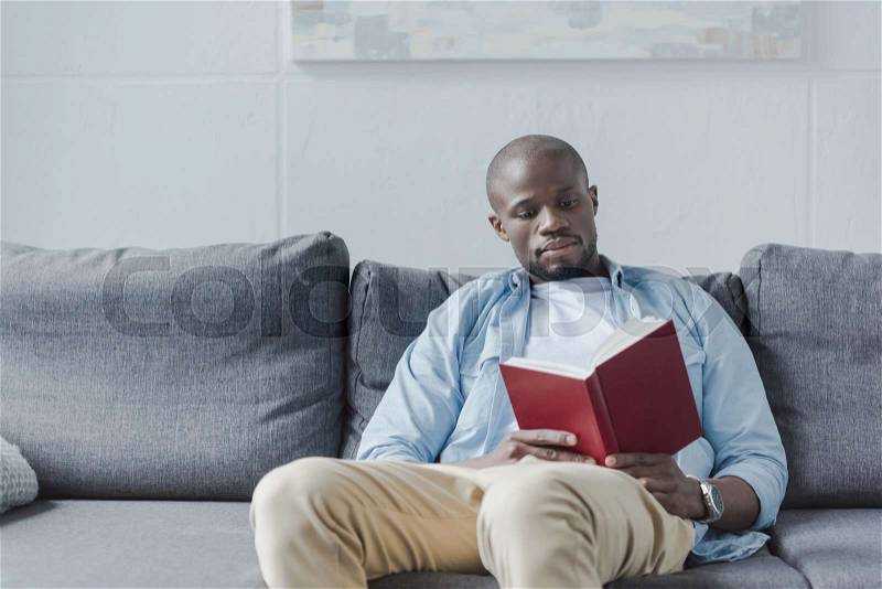 Handsome african american man reading book at home, stock photo
