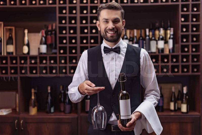 Portrait of handsome sommelier holding bottle of wine and glass in cellar, stock photo