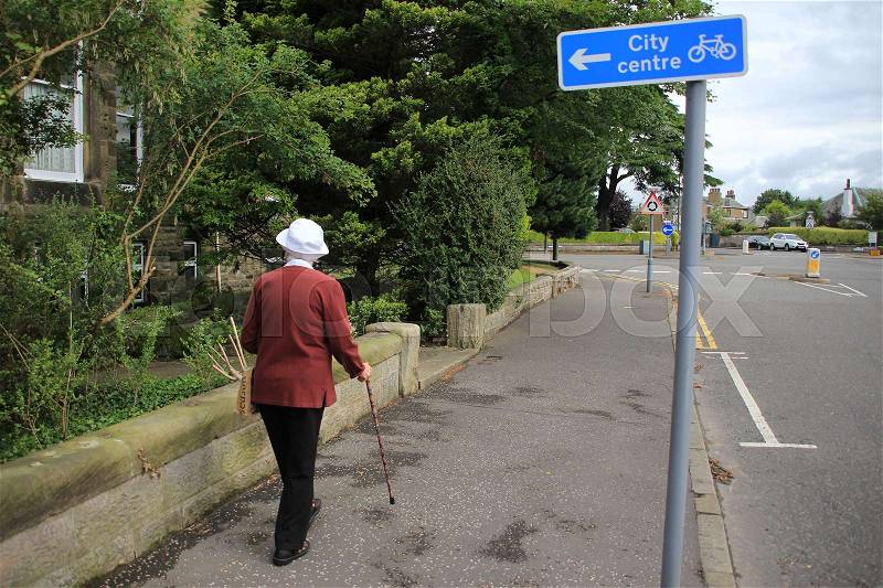 The retired woman with a walking stick is walking in the residential area of the village Stirling in Scotland in the summer, stock photo