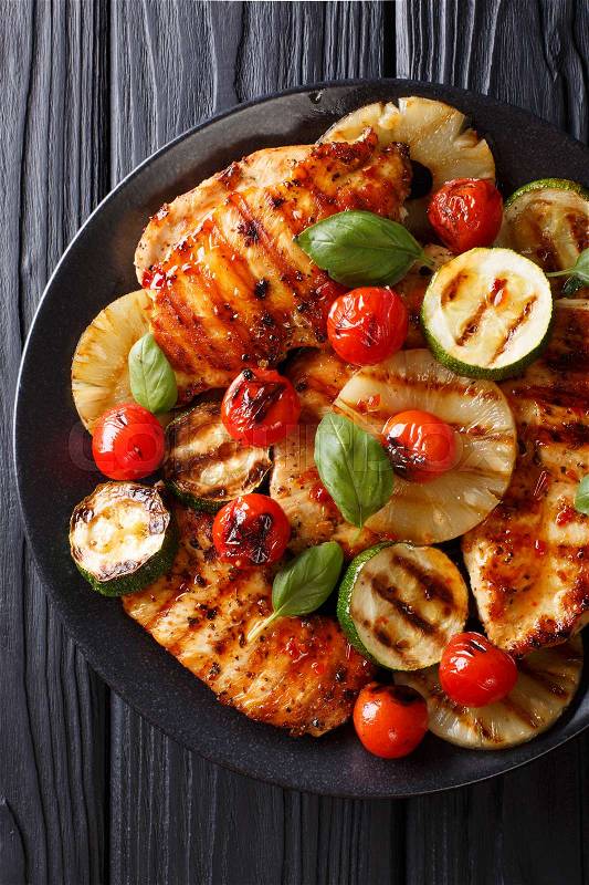 Chicken fillet grilled with pineapple, zucchini and tomatoes in a spicy sauce close-up on a plate. Vertical view from above , stock photo