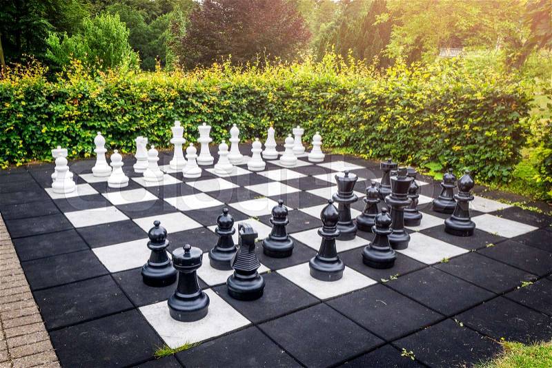 Large outdoor chess game on a garden terrace in a yard with a green hedge and sunshine, stock photo