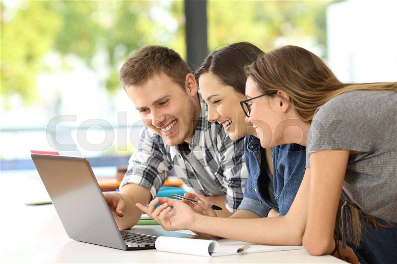 Three happy students learning together on line with a laptop in a classroom, stock photo