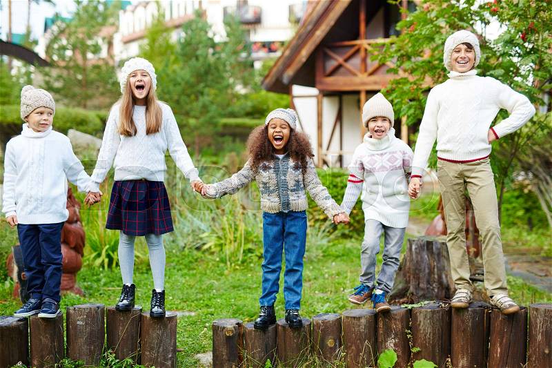 Happy children of various ages having fun playing outdoors in green countryside, standing in row on wooden posts holding hands dressed in similar white knit clothes, stock photo