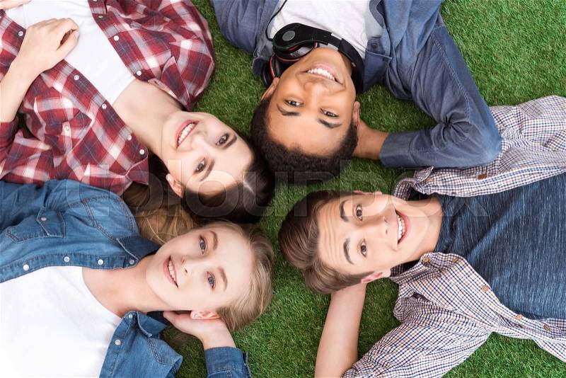 Overhead view of multiethnic group of smiling teenagers lying on green lawn and looking at camera, stock photo