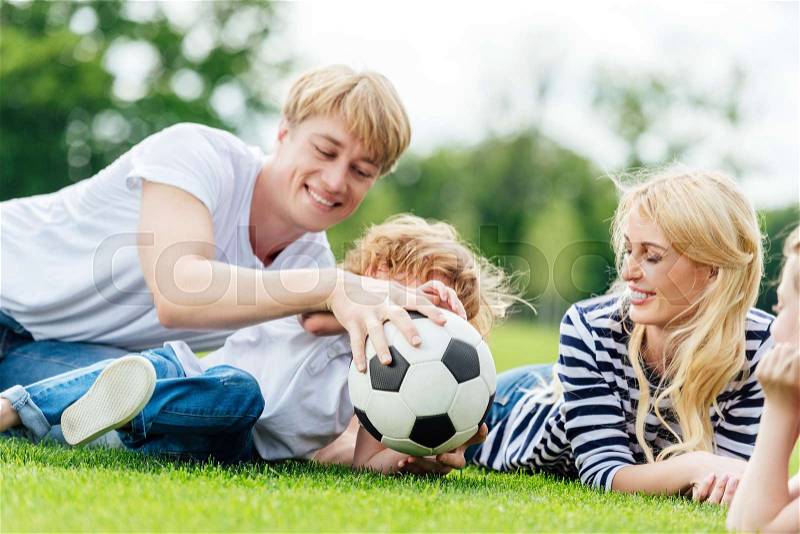 Happy family with two kids having fun with soccer ball while lying on green meadow, stock photo