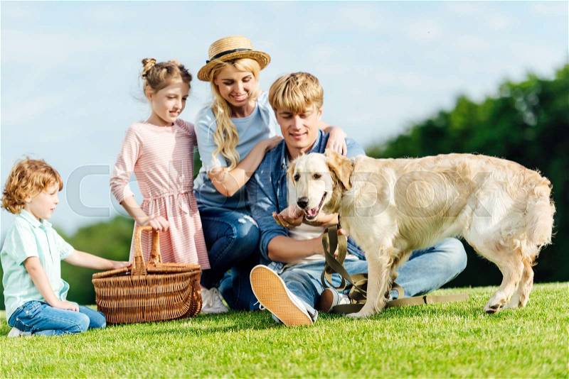 Happy young family with dog and picnic basket resting on green meadow in park, stock photo