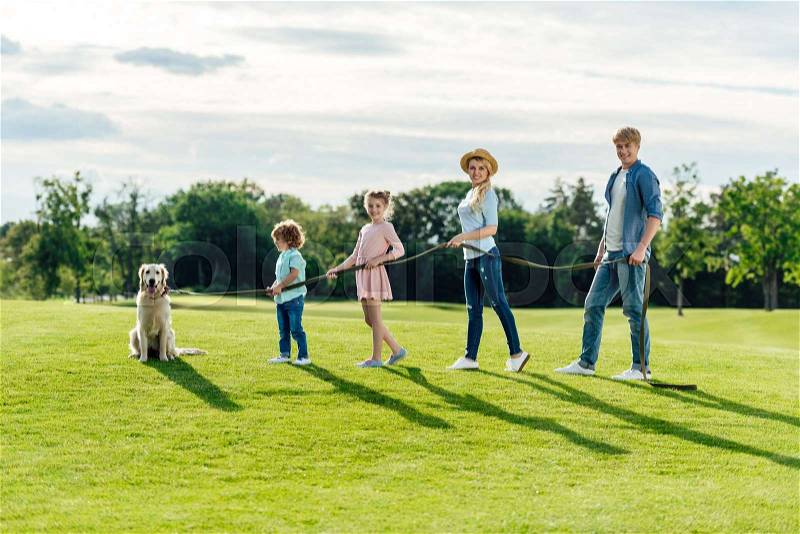 Side view of happy young family with pet walking on green meadow at park, stock photo