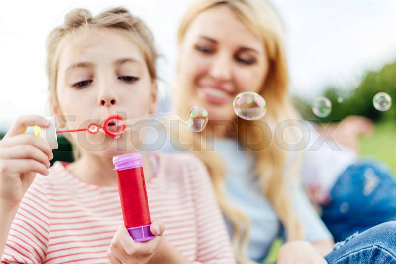 Close-up view of happy mother and daughter blowing soap bubbles in park, stock photo