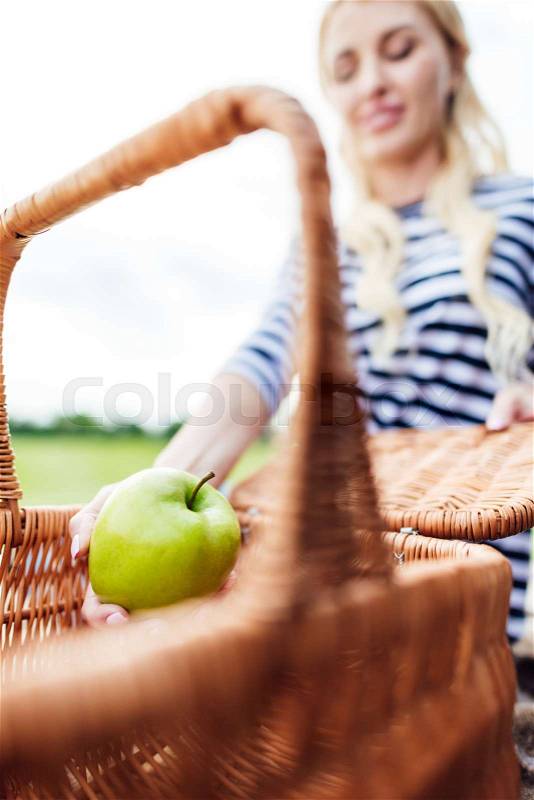Close-up view of young woman holding green apple from picnic basket at park, stock photo