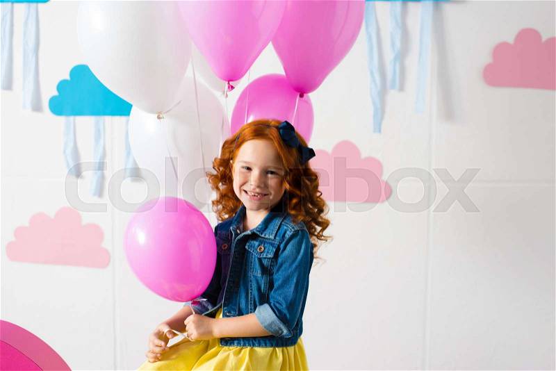 Portrait of adorable red haired girl holding pink balloon and smiling at camera at birthday party, stock photo