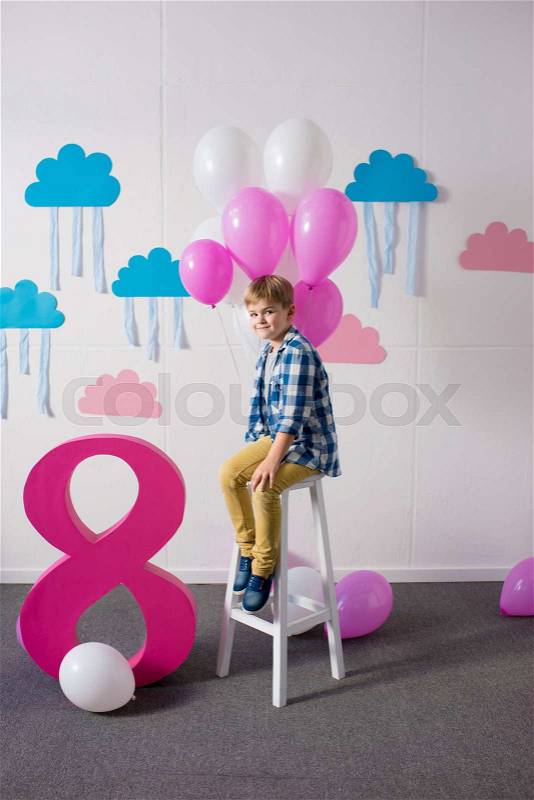 Happy little boy sitting on stool with balloons and smiling at camera at birthday party , stock photo