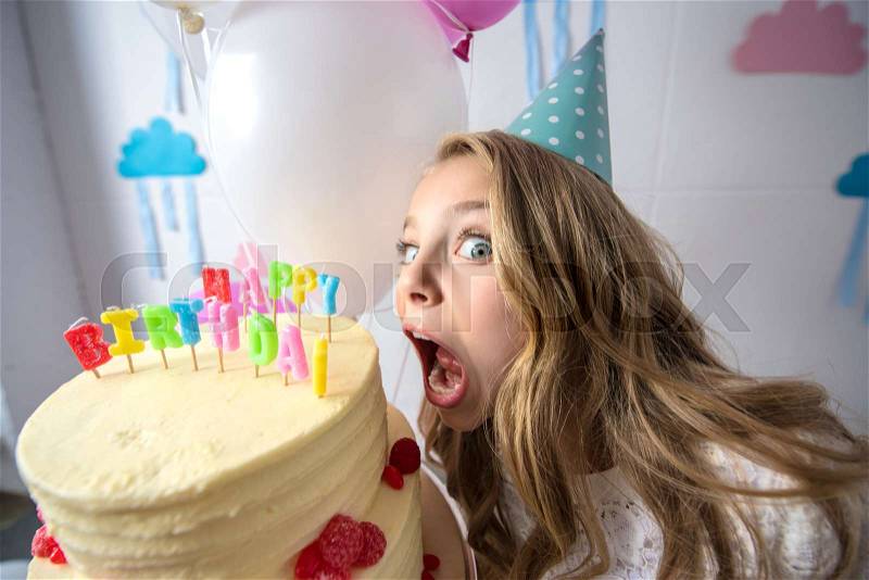 Cute little girl in party cap biting birthday cake and looking at camera, stock photo