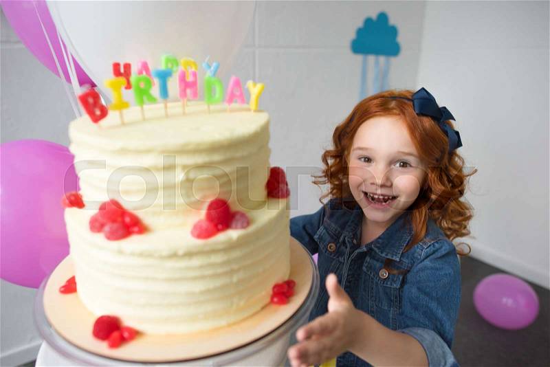 Happy red haired girl reaching to birthday cake and smiling at camera, stock photo