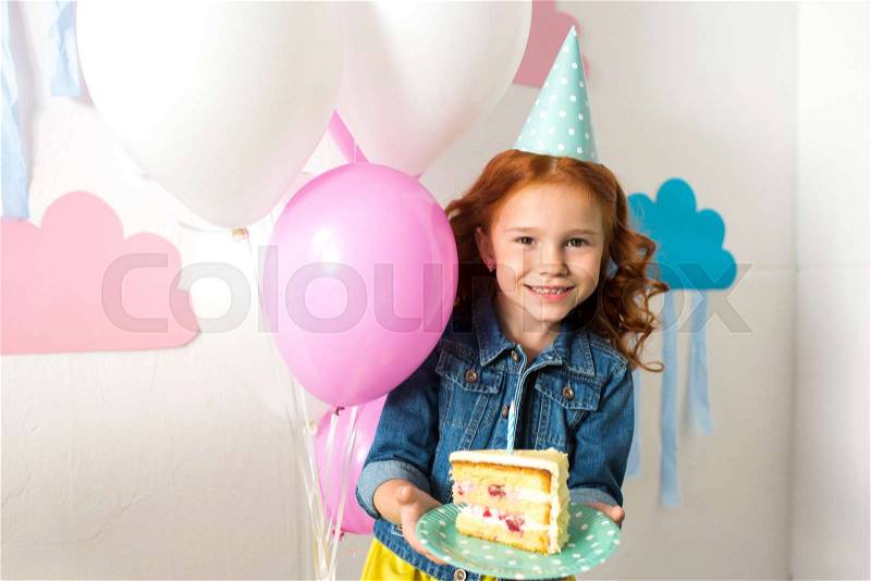 Adorable redhead girl on party hat holding birthday cake and smiling at camera, stock photo