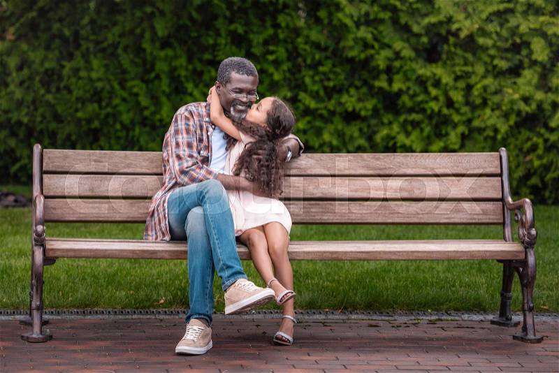 Adorable african american girl hugging and kissing her grandfather while sitting on bench in park , stock photo