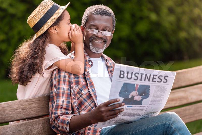 African american girl whispering to her grandfather while he reading business newspaper on bench , stock photo