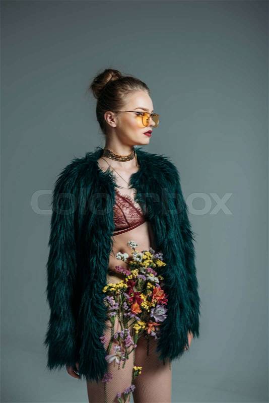 Young attractive model posing in floral skirt, lace bra and green fur coat for fashion shoot, isolated on grey, stock photo