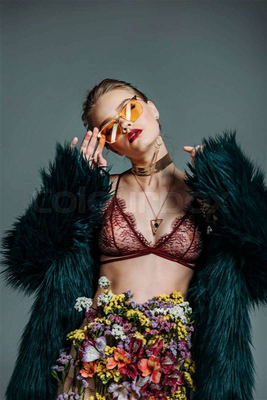Young sensual beautiful girl posing in orange sunglasses, floral skirt, lace bra and green fur coat for fashion shoot, isolated on grey, stock photo