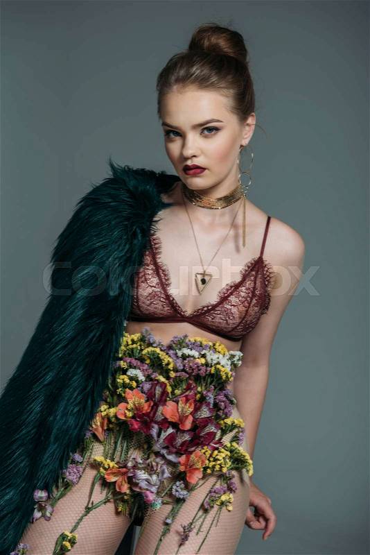 Young sensual stylish model posing in floral skirt, lace bra and green fur coat for fashion shoot on grey, stock photo