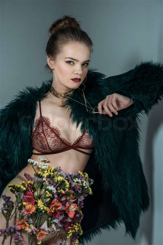 Young sensual beautiful model posing in floral skirt, lace bra and green fur coat for fashion shoot, isolated on grey, stock photo