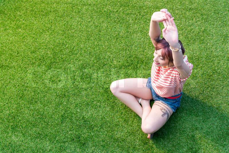 High angle view of smiling young woman stretching hands while sitting on green grass, stock photo