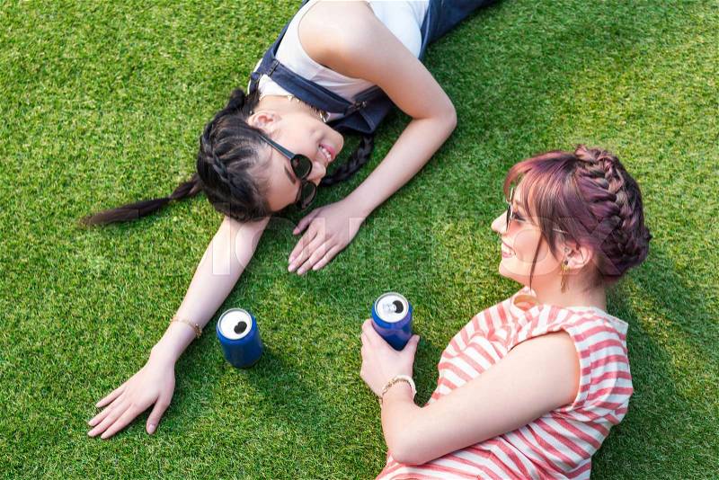 High angle view of multiethnic girls with soda cans smiling each other while lying on lawn, stock photo