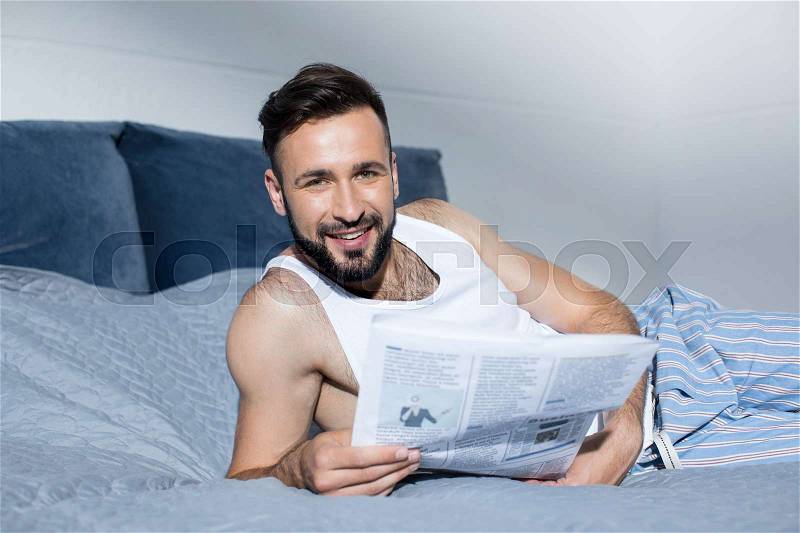 Handsome bearded man reading newspaper and smiling at camera while lying in bed, stock photo